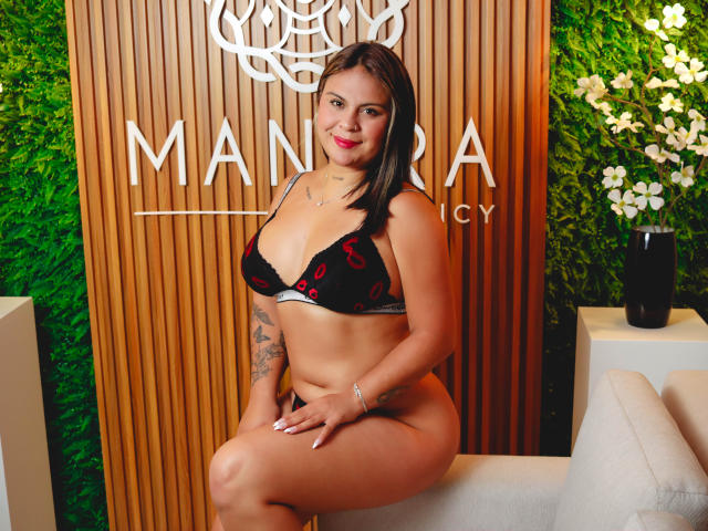 CamillaMoretti with medium tits and a curvy ass, wearing black lace bra and panties with red lipstick prints, sitting on a beige chair, smiling at the camera.