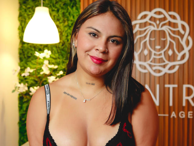CamillaMoretti has medium brown skin, straight black hair, and red lipstick. She's wearing a black lace top, showing her tits, with tattoos on her chest and necklace.