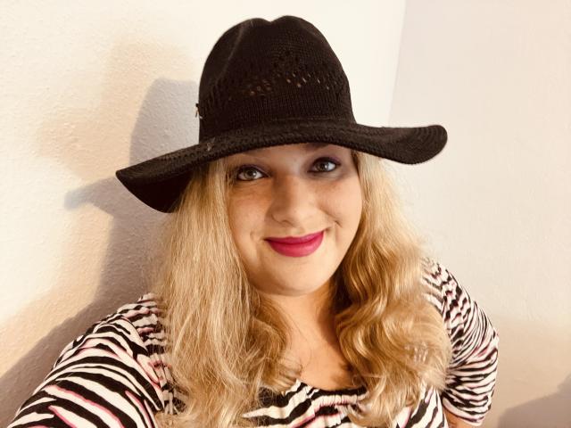 KitsuneAiko, com cabelo loiro ondulado, usando um chapéu de malha preta, uma blusa listrada como zebra e batom rosa, sorrindo para a câmera. Seus seios estão ligeiramente visíveis sob a blusa..