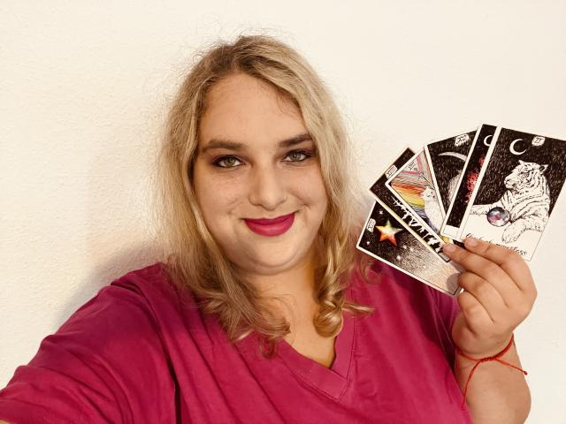 KitsuneAiko, rubia con cabello rizado, lápiz labial rosa, camisa roja, sosteniendo cartas de tarot, sonriendo, fondo de pared blanca, pulsera roja, ligera barriga, selfie casual, segura de sí misma, ambiente relajado..