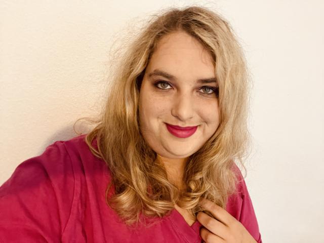 KitsuneAiko, con cabello rubio ondulado, lápiz labial rosa y una camisa roja, sonríe a la cámara, sosteniendo el cuello de su camisa. Sus senos son de tamaño promedio y tiene piel clara..