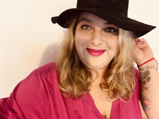 KitsuneAiko met blond, golvend haar, een zwarte hoed, een roze shirt, rode lippenstift, lachend, met een zichtbare tatoeage op de rechterarm, die een hoed vasthoudt, een rode armband, en een witte muur op de achtergrond..