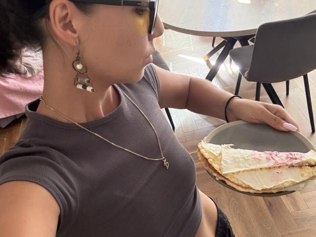 Jeannie Dobransky, con i capelli scuri raccolti in una coda di cavallo, indossa occhiali e una maglietta grigia, tiene un piatto con una torta parzialmente mangiata e sbriciolata, e la sua mano è appoggiata su una sedia..