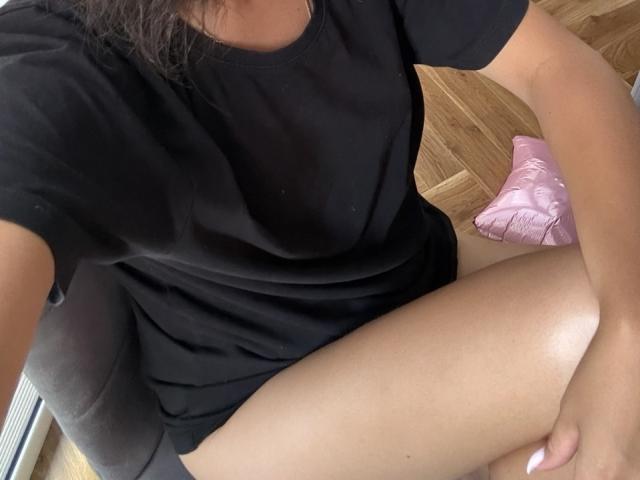 JeannieDobransky's black t-shirt, her bare thighs, and a pink pillow on a wooden floor. She's sitting with her right arm resting on her knee, head partially out of frame.