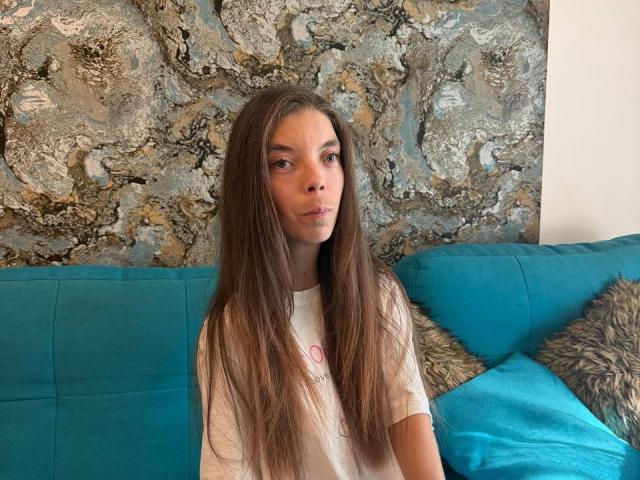 OlishaSpace, avec de longs cheveux bruns, des yeux bleus et un t-shirt blanc, est assise sur un canapé turquoise avec des coussins moelleux, et a l'air légèrement ennuyée. Un mur avec un motif de marbre se trouve derrière elle..