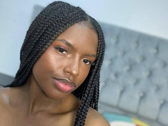 EllysRose has dark brown skin, long black braids, and glossy pink lips. She's topless, showing her shoulders. Her makeup includes subtle eyeshadow and defined eyebrows.