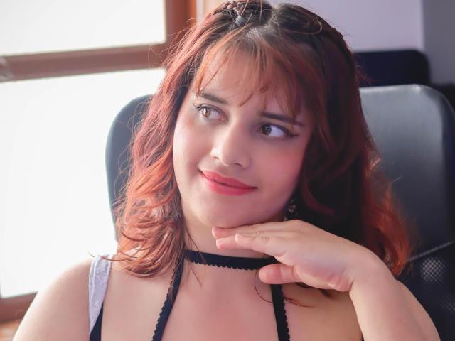 VivianFranco with red, wavy hair, wearing a black choker, smiling softly, her hand gently touching her chin, in a bright, sunlit room. She has cute, small tits.