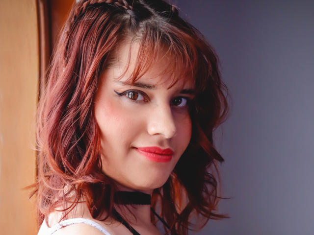 VivianFranco has red-brown wavy hair, black choker, white tank top, red lipstick, and black winged eyeliner, smiling with brown eyes. Her tits are not visible.
