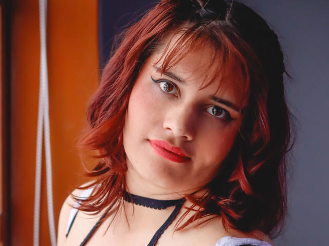 VivianFranco with red hair, black choker, and white top, looking slightly to the side with a subtle smile. Her tits are small and her pussy is not visible.