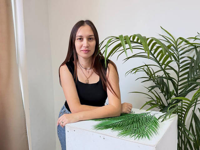 DinaPeich with long brown hair, black tank top, and blue jeans, leaning on a white box with green palm leaves, smiling slightly, wearing a gold necklace.