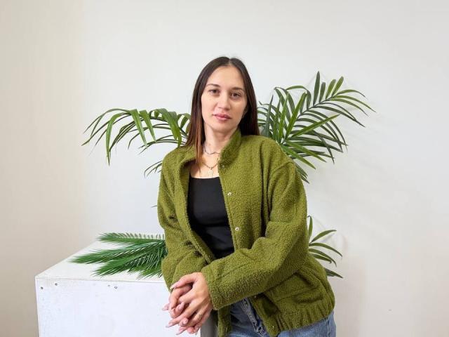 DinaPeich, com cabelo castanho, um cardigã verde felpudo, blusa preta, jeans azuis, mãos juntas, plantas verdes ao fundo, parede branca, sorrindo, fofa, seios médios, pele clara, casual..