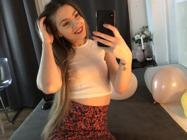 Lesysl with long brown hair, smiling, wearing a white crop top and floral skirt, taking a mirror selfie with a black phone, showing her tits and ass. Balloons in background.