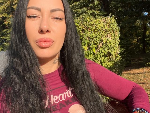 MirandaHott69 with long black hair, tan skin, full lips, and eyebrows, wearing a red long-sleeve shirt, pouting slightly, sunlight highlighting her face.