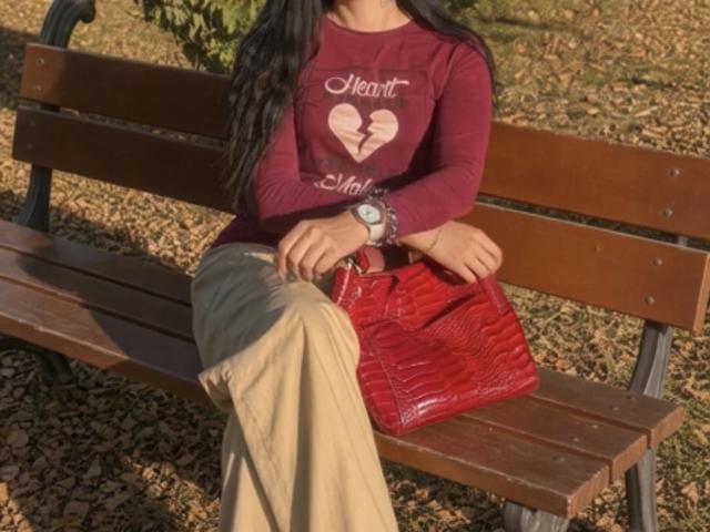 MirandaHott69 sitting on a wooden bench, wearing a red heart shirt, beige pants, red croc handbag, and a green wristwatch, with long black hair.