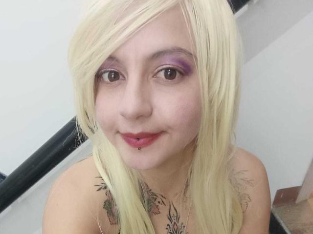 ArianaBlonde with blonde hair, purple eye makeup, and a nose piercing, smiling with a lip ring, showing tattoos on her chest, sitting with a black railing behind.