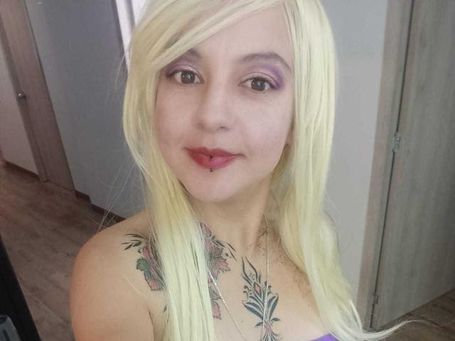 ArianaBlonde with long blonde hair, pink eyeshadow, red lipstick, pierced lower lip, tattoos on chest, looking at camera, smiling, topless, in a simple room.
