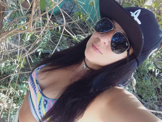 SexyMaxine with long black hair, wearing a black cap with an "A", sunglasses, and a colorful bikini top, showing her tits. She's outdoors with greenery around.
