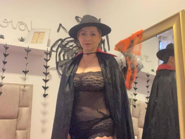 GianineLarose wearing a black witch hat, sheer black lace lingerie, and a black cape, showing her tits and ass, standing in a mirror's reflection.