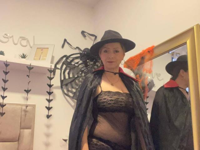 GianineLarose in a black witch outfit with a hat, black lace lingerie, and a black cape, standing in front of a mirror, showing her tits and reflection.