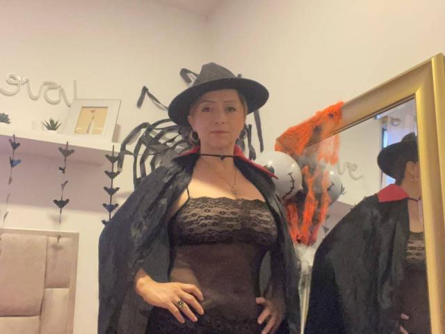 GianineLarose with short black hair, wearing a black lace strapless top, black cape with red trim, and black witch hat, standing in front of a mirror, hands on hips.