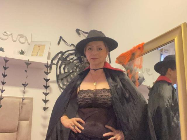 GianineLarose in a black witch hat and lace bustier, black cape, red collar, standing with hands on belly, reflection in mirror, gold frame, looking at camera, medium breasts.