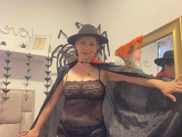 GianineLarose wearing a witch hat and black lace lingerie with a sheer black cape, exposing her tits, in a mirror reflection, with a cute, slightly smiling face.