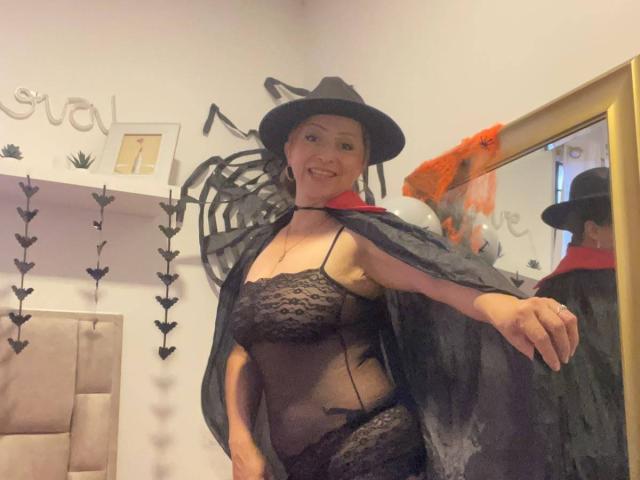 GianineLarose with black witch hat, black lace lingerie, and sheer black cape, exposing her tits and pussy under the see-through fabric, smiling in a mirror.