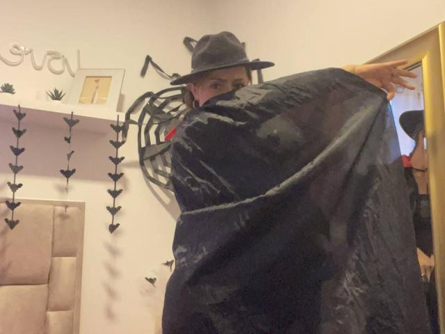 GianineLarose wearing a black witch hat and cape, arms spread, showing a small butt, in a dimly lit room with heart decorations and a padded headboard.