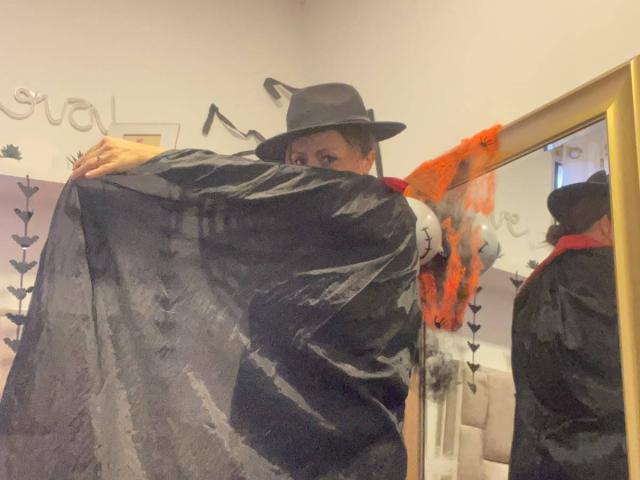 GianineLarose in a black witch hat and sheer black robe, back to camera, arms up, reflecting in a mirror, orange lace on right, white walls, yellow mirror frame.