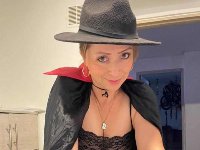 GianineLarose in a black witch hat, black lace top, red collar, and black cape, with a necklace and hoop earrings, smiling with her tits slightly showing.
