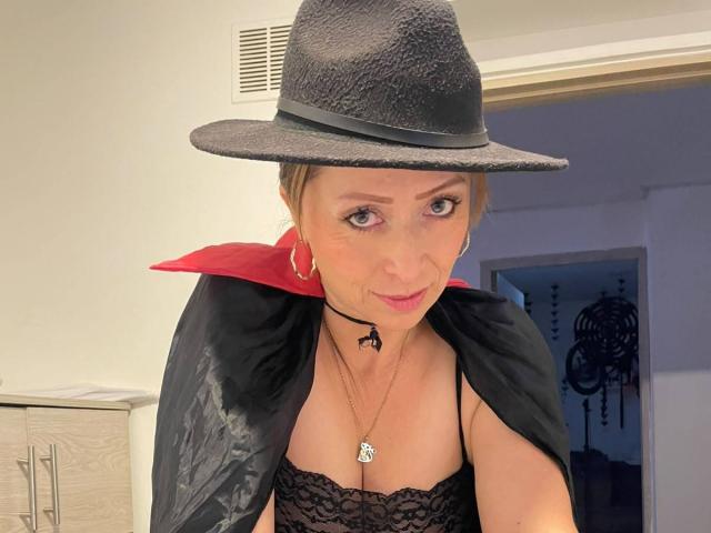 GianineLarose with black witch hat, red collar, black lace top, and necklace, showing her tits. Blonde, hoop earrings, slight smile, in a dimly lit room.