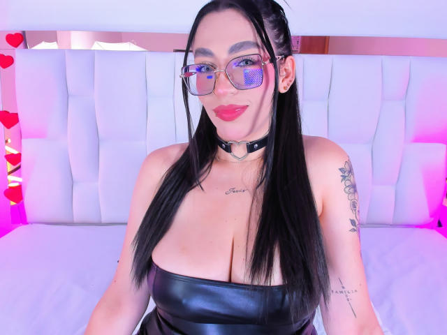 CamilleJuicyHot with long black hair, glasses, red lipstick, black leather strapless top, choker, tattoos on arms, big boobs, pink and purple lighting, smiling.