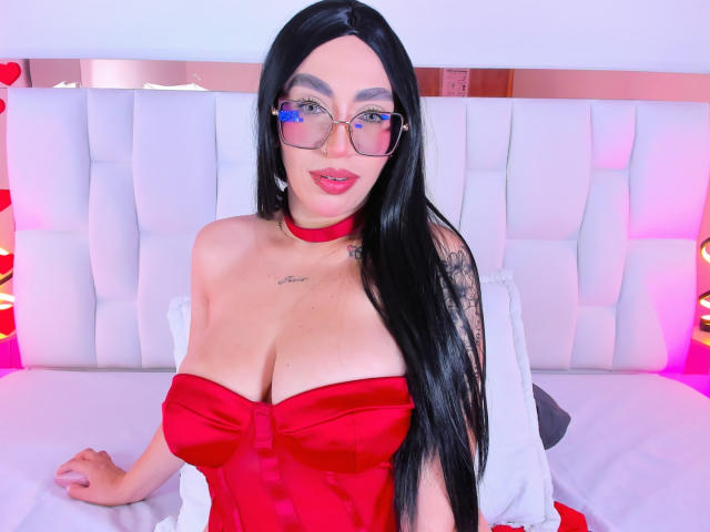 CamilleJuicyHot with long black hair, glasses, red choker, red strapless dress, big tits, red lipstick, sitting on bed, white headboard, pink and purple lighting.