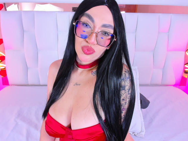 CamilleJuicyHot with long black hair, glasses, red choker, and red dress showing big tits, blue lipstick, and tattoo on right arm, sitting on a white bed.