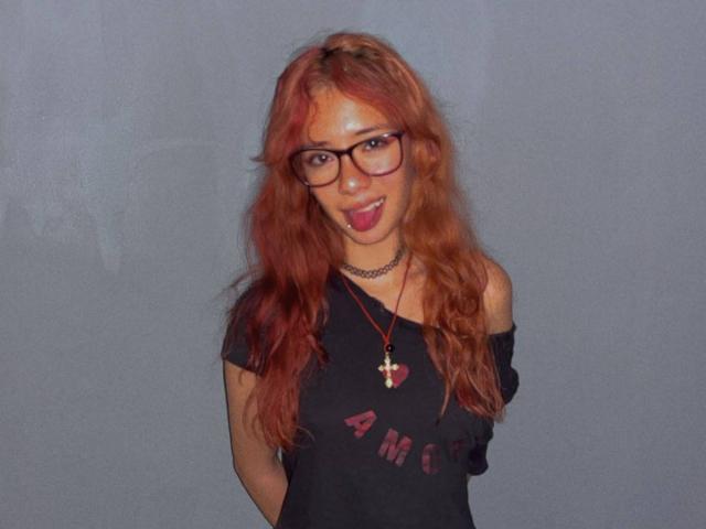 SuriEverly with long red hair, glasses, black off-shoulder shirt, choker, and cross necklace, sticking out tongue, arms behind back, looking cute and nerdy.
