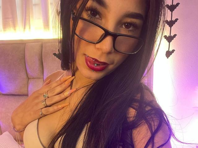 VioletaVelvet, con lunghi capelli neri, occhiali e rossetto rosso, mostra il seno con una mano, indossa anelli alle unghie e ha un piercing alla lingua, in una stanza con una luce soffusa..