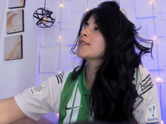 NinaHarpper with long black hair, wearing a green and white soccer jersey, slightly open mouth, looking to the side, soft purple light, and a black geometric hanging light.