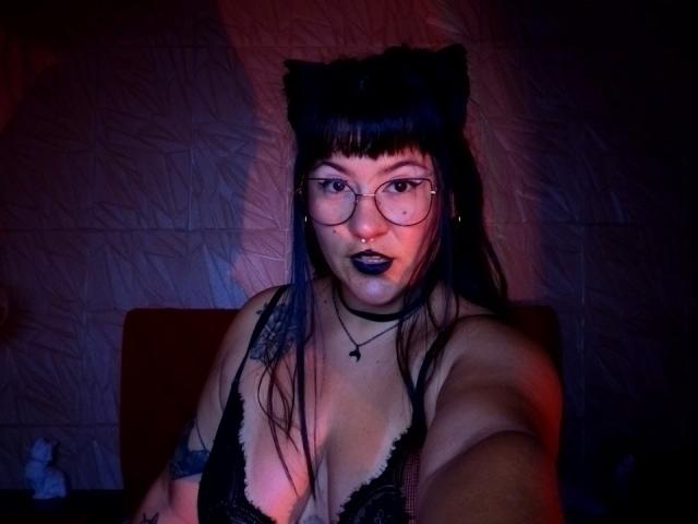 NaniSky with cat ears, glasses, black lipstick, and black lace bra, showing off her tits and tattoos, taking a selfie with red and blue lighting.