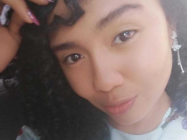 KirasPell's pouty face with curly black hair, brown eyes, and light brown skin, slightly puffed pink lips, and a hand with pink nails touching her head.
