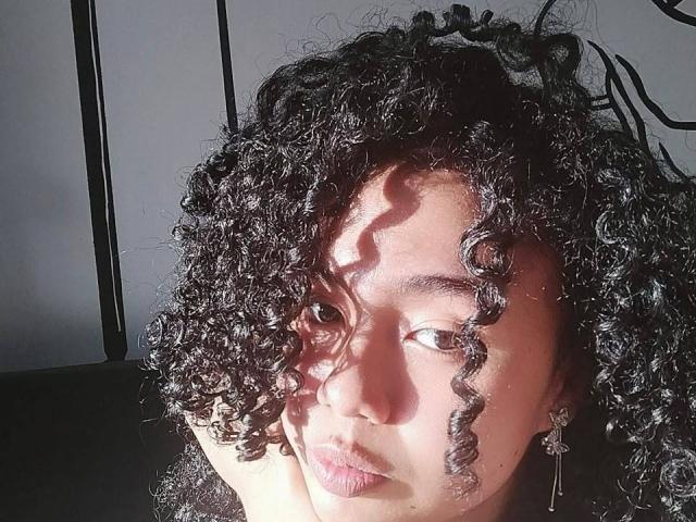 KirasPell's curly black hair casts shadows on her face, sunlight highlighting her dark eyes and pink lips. She wears dangling earrings, looking slightly to the side with a serious expression.