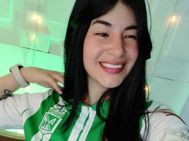 NinaHarpper with long black hair, green and white striped soccer shirt, smiling, pink lipstick, wrist tattoo, smartwatch, green-lit background.