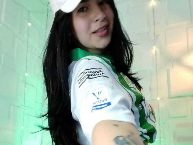 NinaHarpper with long black hair, wearing a white cap, green and white soccer jersey, smiling at the camera, showing a tattoo on her right arm, and a cute face.