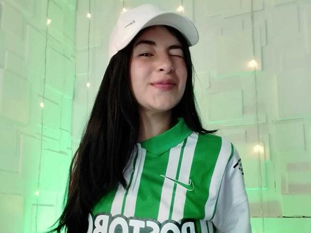 NinaHarpper with long black hair, wearing a white cap and green and white soccer jersey, smiling with one eye closed, showing her cute, small tits.