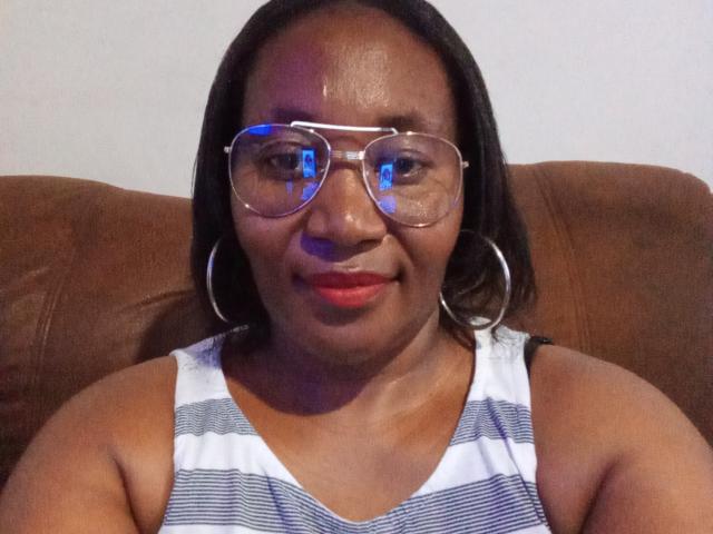 CavyQueenForU heeft een donkere huid, draagt een bril met blauwe reflecties, een gestreept wit tanktopje en rode lippenstift, en zit op een bruine leren bank..