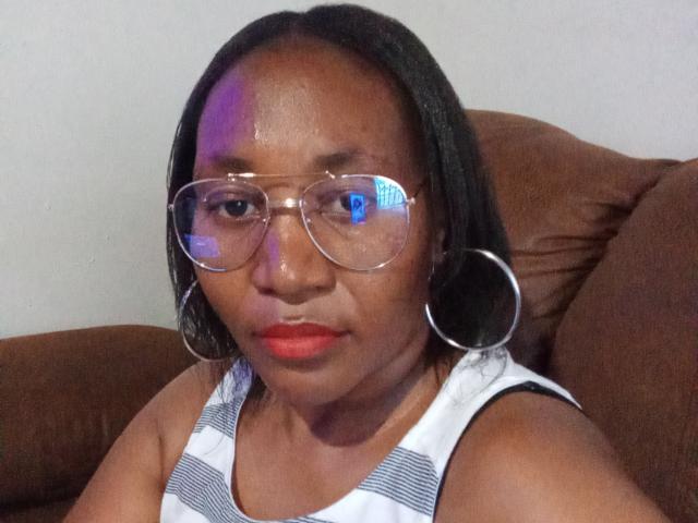 CavyQueenForU has dark skin, black straight hair, red lipstick, clear glasses, and large hoop earrings. She's wearing a white and gray striped tank top.