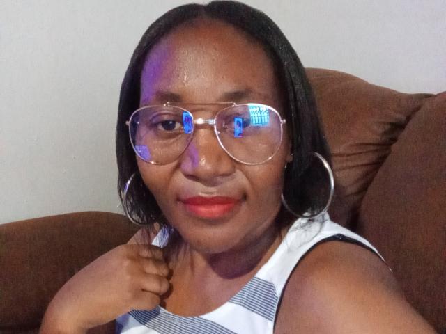 CavyQueenForU con piel oscura, cabello negro de largo medio, usando gafas transparentes que reflejan luz azul, una blusa blanca a rayas sin mangas, lápiz labial rojo, grandes aros, sentada en un sofá marrón..