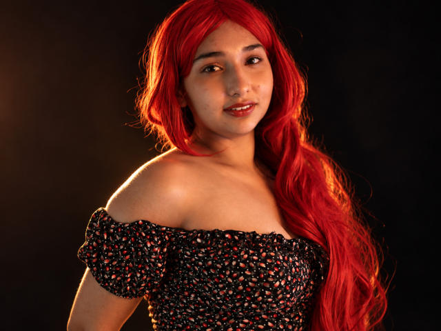EleanorLamberth with long red hair, off-shoulder black floral dress, smiling at the camera, showing her bare shoulders and soft skin, against a dark background.