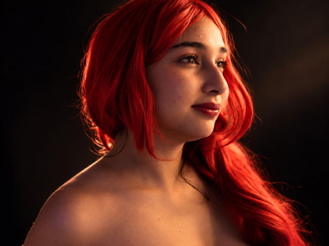 Eleanor Lamberth tem cabelos ruivos vibrantes, pele clara e um leve sorriso. Ela está sem blusa, mostrando os ombros nus. Seus olhos castanhos estão voltados para o lado, iluminados por uma luz quente..