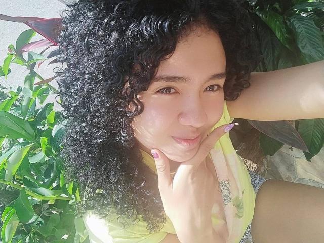 KirasPell with curly black hair, light brown skin, and purple nails, slightly pouting, holding her chin, wearing a yellow top, and sitting amid green plants.