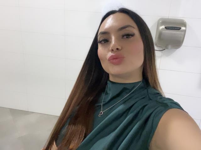GalaMalkova with long brown hair, green sleeveless top, and pink pout, wearing a silver necklace, looking at the camera in a bathroom.
